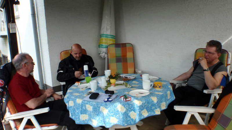 Drei Männer sitzen um einen mit blauem Tischtuch bedeckten Tisch auf einer Terrasse.