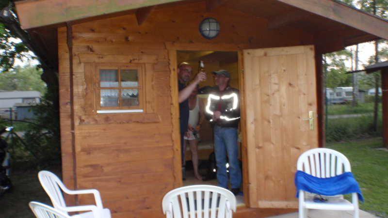 Zwei Männer stehen in der offenen Tür einer kleinen Holzhütte und stoßen mit Getränken an, davor stehen weiße Kunststoffstühle.