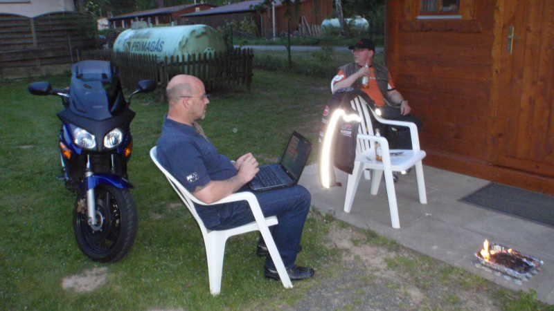 Zwei Männer sitzen auf weißen Plastikstühlen vor einer Holzhütte neben einem Motorrad und einem kleinen Feuer im Freien