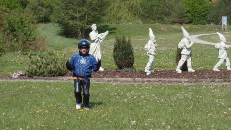 Kind mit Helm und Schutzkleidung läuft über eine Wiese vor weißen Gartenzwerg- und Frauenstatuen