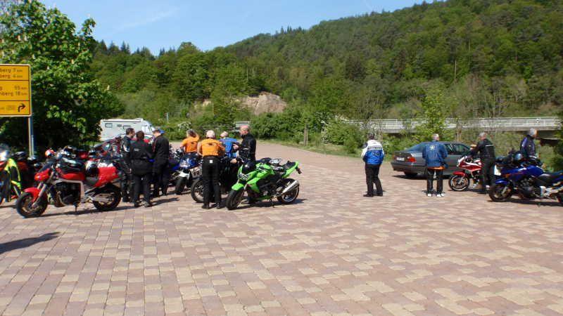 Gruppe von Motorradfahrern mit Motorrädern auf gepflastertem Platz vor bewaldeten Hügeln