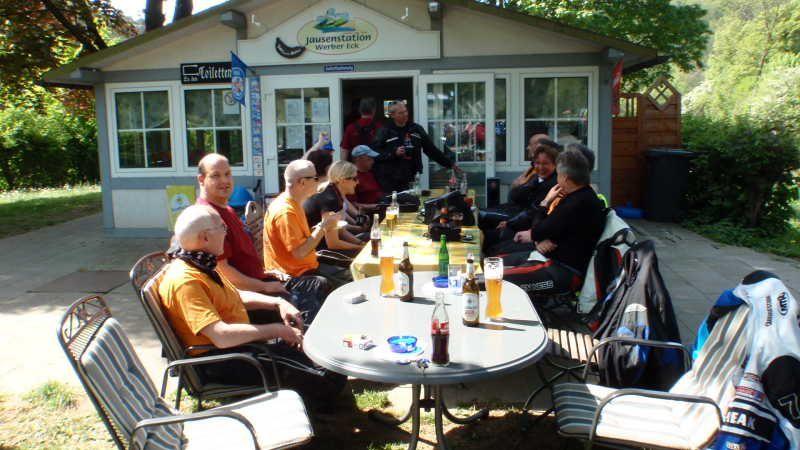 Gruppe von Menschen sitzt draußen an einem langen Tisch vor einem Imbissgebäude