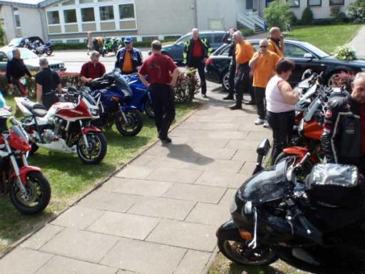 Mehrere Menschen stehen auf einem Gehweg neben einer Reihe geparkter Motorräder in einem Wohngebiet.