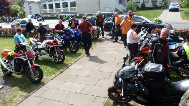 Mehrere Menschen stehen auf einem Gehweg neben einer Reihe geparkter Motorräder in einem Wohngebiet.