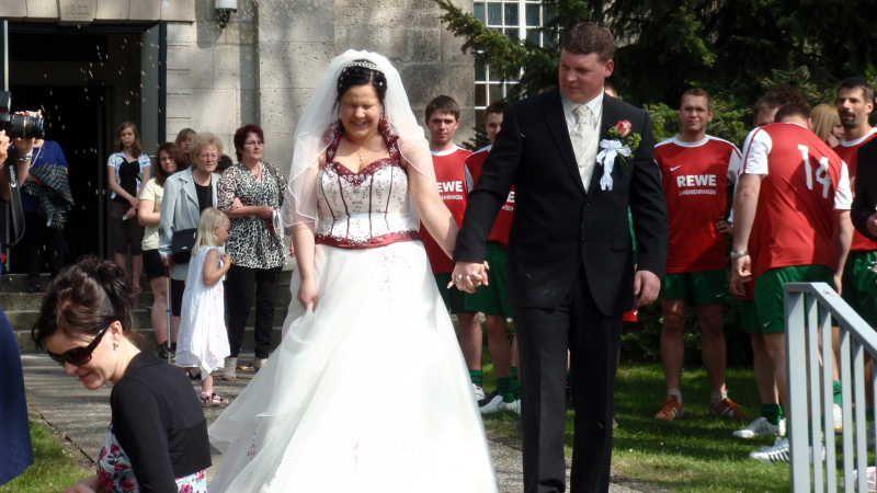 Braut und Bräutigam in Hochzeitskleidung gehen Hand in Hand vor einer Gruppe von Menschen, darunter Fußballer in roten Trikots.