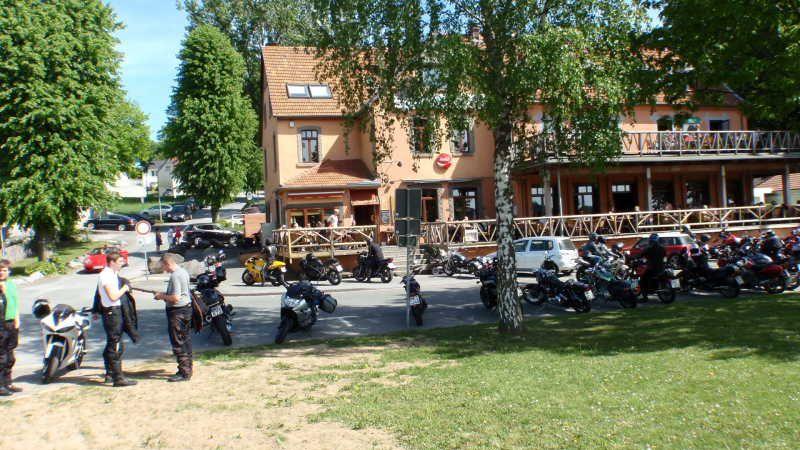 Mehrere Motorräder und Menschen vor einem zweistöckigen Gebäude mit Terrasse und Bäumen im Hintergrund