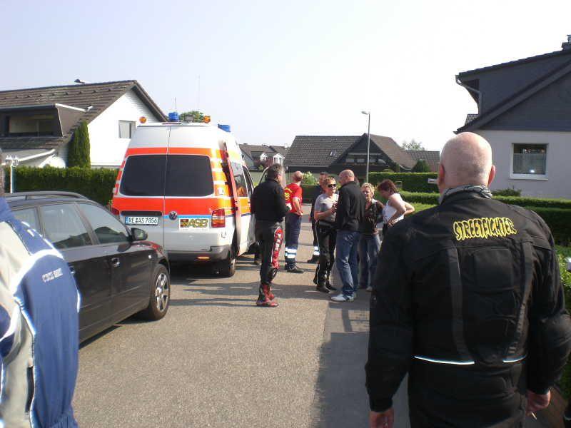 Gruppe von Menschen steht auf einer Straße neben einem Rettungswagen und mehreren Autos in einem Wohngebiet