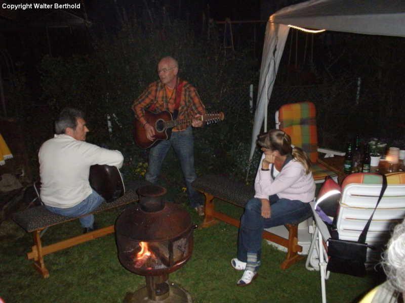 Drei Personen im Garten bei Nacht mit zwei Gitarren und einem Feuerofen