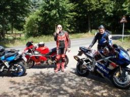 Zwei Motorradfahrer in Schutzkleidung stehen neben ihren Motorrädern auf einer Parkfläche im Grünen.