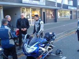 Fünf Motorradfahrer in Schutzkleidung stehen neben parkenden Motorrädern auf einem Bürgersteig vor Geschäftsgebäuden.