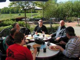 Fünf Personen sitzen um zwei runde Tische im Freien und essen gemeinsam.