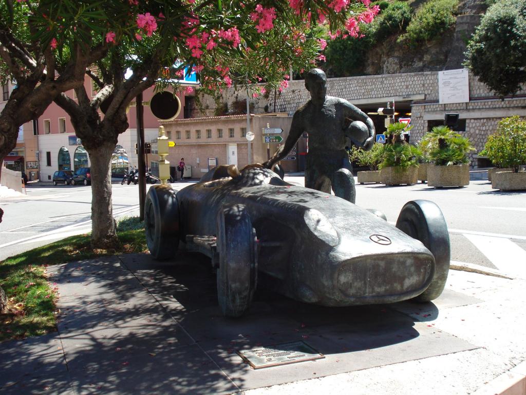 Bronzestatue eines Rennwagens mit einem Fahrer, der einen Helm hält, unter einem blühenden Baum an einer Straßenecke
