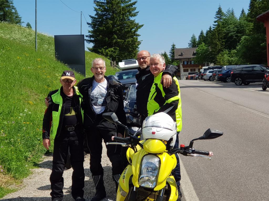 Vier Männer in Motorradbekleidung stehen neben einer gelben Motorrad auf einer Straße neben einer Wiese und Parkplätzen.
