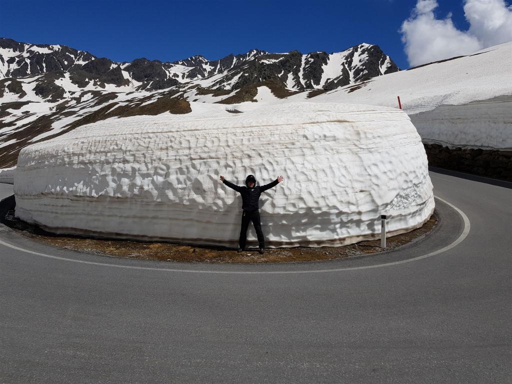 Person steht vor einer hohen Schneewand an einer Bergstraße mit schneebedeckten Bergen im Hintergrund