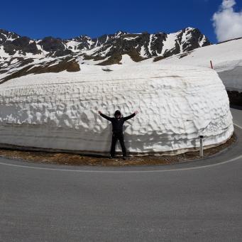 Person steht vor einer hohen Schneewand an einer Bergstraße mit schneebedeckten Bergen im Hintergrund
