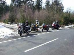 Fünf Motorräder am Straßenrand mit drei Personen in Motorradbekleidung neben einem Wald