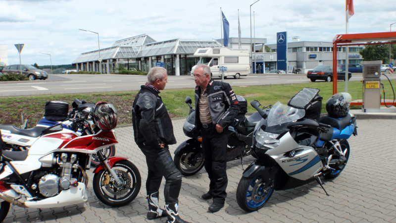 Zwei Männer in Motorradbekleidung stehen auf gepflastertem Parkplatz neben drei Motorrädern und unterhalten sich.