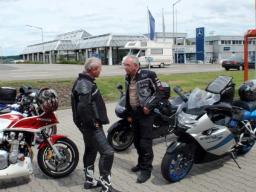 Zwei Männer in Motorradbekleidung stehen auf gepflastertem Parkplatz neben drei Motorrädern und unterhalten sich.