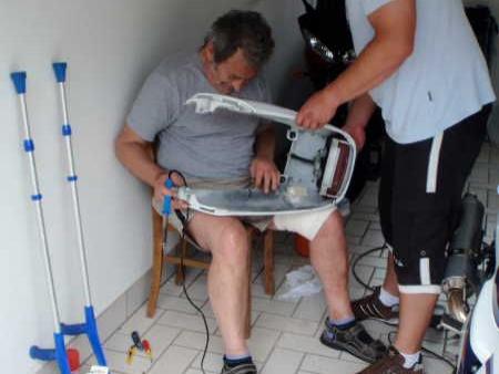 Zwei Männer in einer Garage reparieren ein Autoteil auf einem Stuhl sitzend