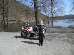 Frau in Motorradbekleidung vor einem Motorrad an einem See mit Bäumen