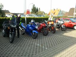 Mehrere farbige Motorräder und zwei Personen auf einem Parkplatz vor Häusern