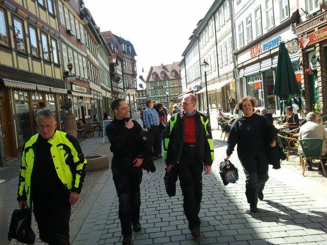 Vier Personen in Motorradkleidung gehen eine gepflasterte Straße in einer Altstadt entlang.