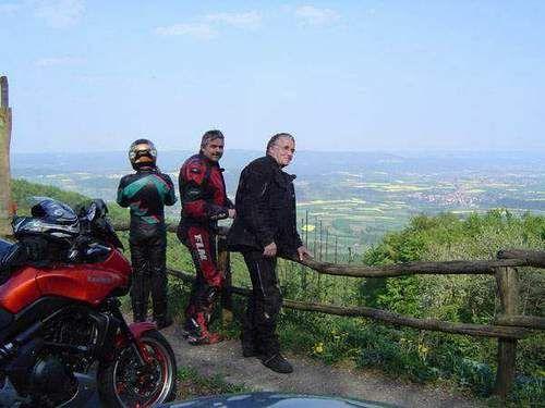 Drei Motorradfahrer stehen an einer Holzbrüstung mit Blick auf ein Tal.