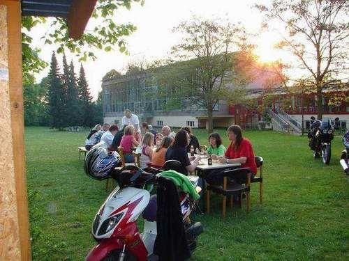 Gruppe von Menschen sitzt an Tischen auf einer Wiese im Freien bei Sonnenuntergang, im Vordergrund ein roter Motorroller