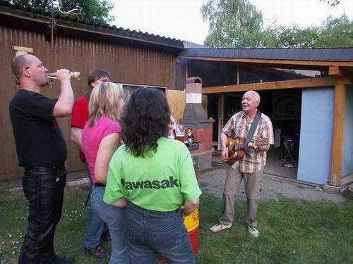 Mehrere Personen stehen draußen vor einem Holzhaus, ein Mann spielt Gitarre und die anderen hören zu
