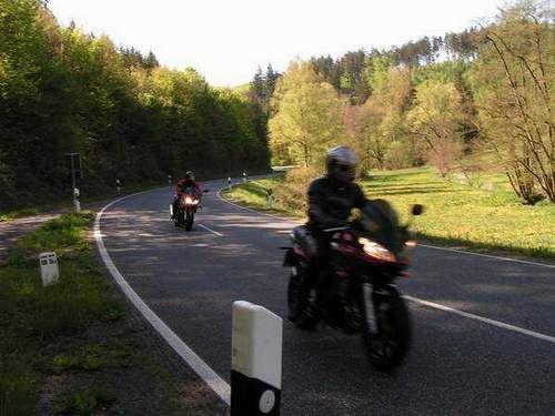 Zwei Motorradfahrer auf einer kurvigen Landstraße mit Wald und Wiese im Hintergrund