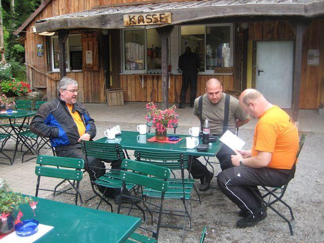 Drei Männer sitzen an einem grünen Gartentisch vor einer Holzhütte mit Kassenschild