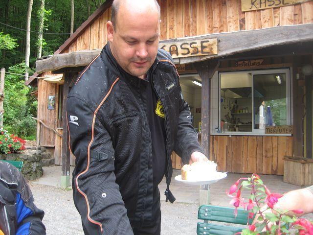 Mann hält einen Teller mit Kuchen vor einem Holzgebäude mit Kassenschild