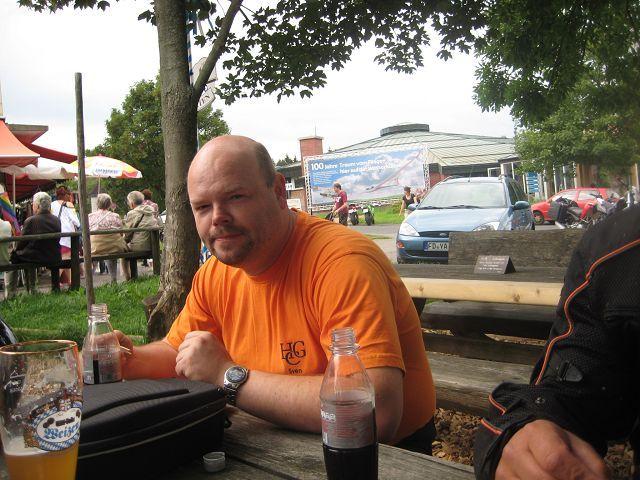 Mann sitzt im orangenen T-Shirt an einem Holztisch im Freien mit Getränken vor sich