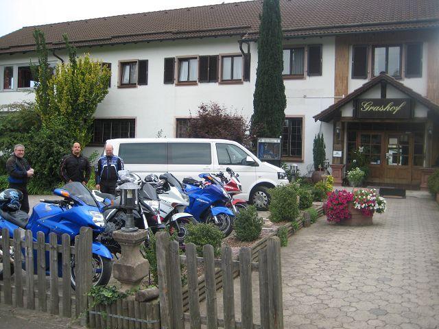 Drei Männer stehen neben mehreren Motorrädern und einem weißen Van vor einem Gasthof.