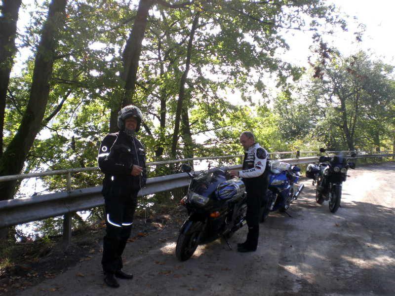 Zwei Männer in Motorradkleidung neben geparkten Motorrädern an einer Straße mit Geländer und Bäumen