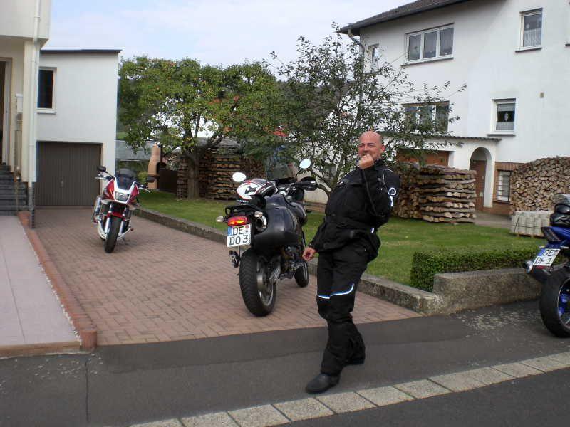 Mann in schwarzer Motorradkleidung vor Motorrädern vor einem Wohnhaus