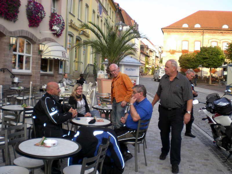 Mehrere Personen sitzen und stehen an einem Straßencafé in einer historischen Innenstadt mit Blumen an der Fassade.
