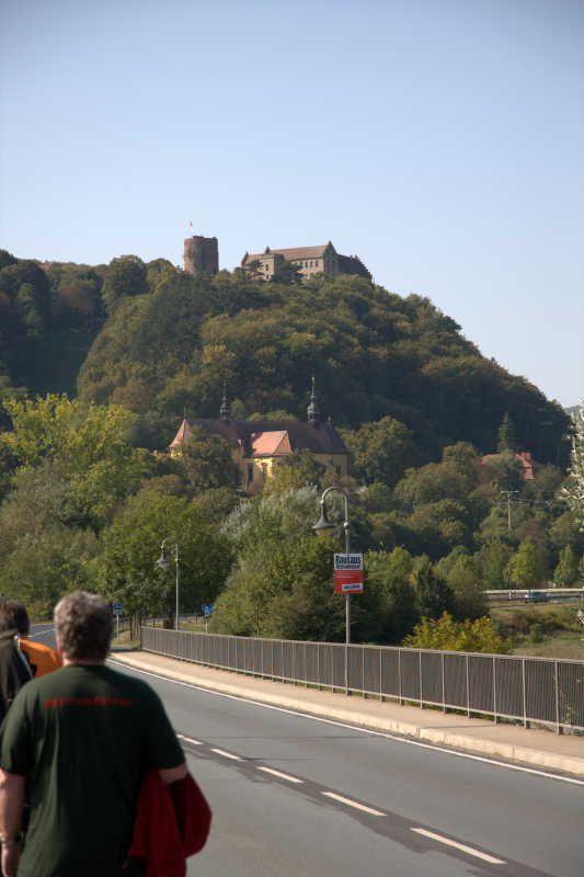 Berg mit bewaldetem Hang, darauf eine Burg und eine Kirche, im Vordergrund eine Straße mit Personen.
