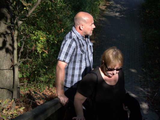 Ein Mann und eine Frau gehen einen schattigen Waldweg hinauf, umgeben von Bäumen und Sträuchern.