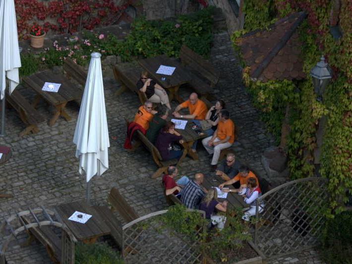 Gruppe von Menschen sitzt an Tischen im Außenbereich eines Restaurants mit gepflastertem Boden und umliegender Bepflanzung
