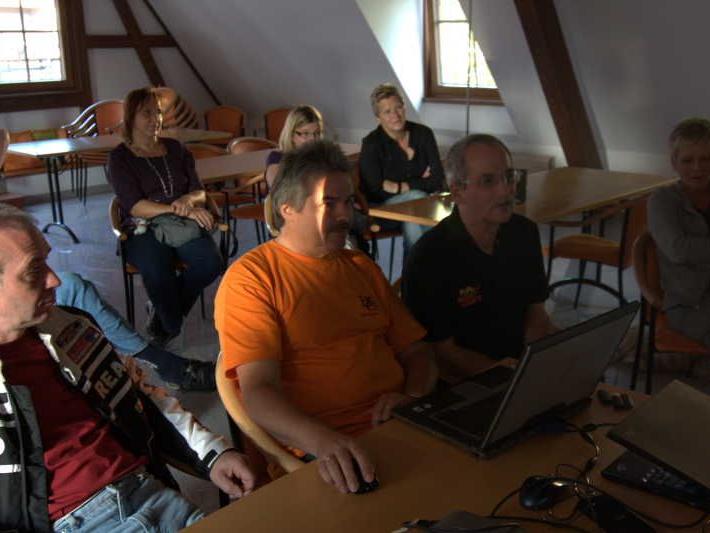 Mehrere Erwachsene sitzen in einem Raum an Tischen und schauen auf einen Laptop.