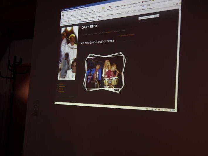An eine Wand projizierte Computerbildschirmansicht einer Webseite über Gary Reck mit Fotos von Personen auf einer Bühne