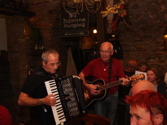 Zwei Männer spielen live Musik mit Akkordeon und Gitarre in einer rustikalen Kneipe vor sitzenden Gästen.