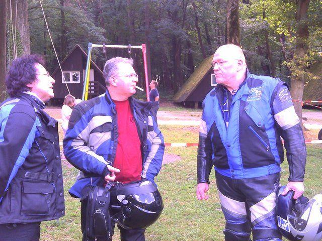 Drei Männer in Motorradkleidung unterhalten sich auf einer Wiese vor einem Wald