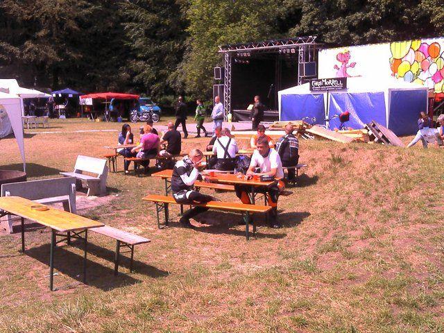 Mehrere Menschen sitzen an Biergartenbänken auf einer Wiese vor einer Bühne bei einem Open-Air-Event.
