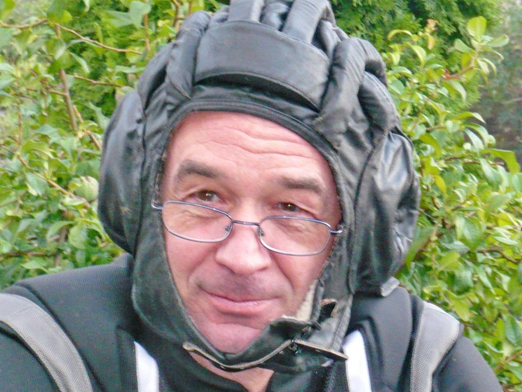 Mann mit Brille trägt einen gepolsterten schwarzen Kopfschutz im Freien vor grünen Pflanzen