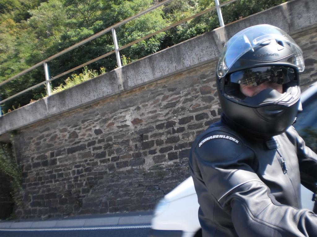 Motorradfahrer mit schwarzem Helm und Lederjacke vor einer Steinmauer und einem vorbeifahrenden weißen Auto