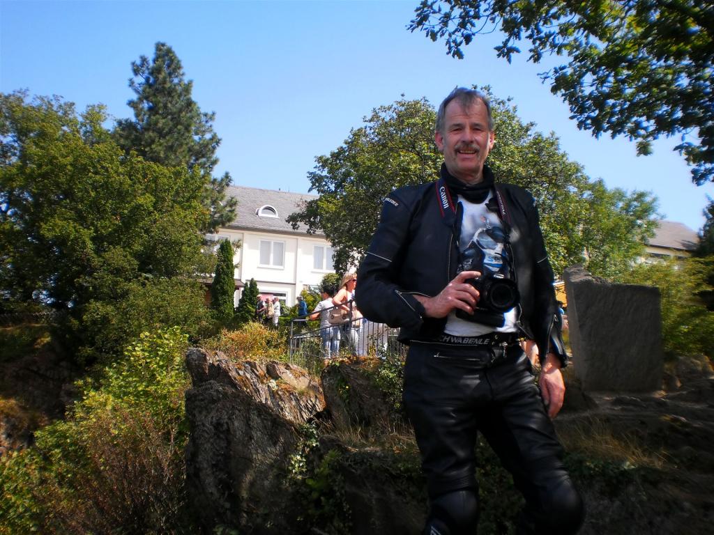 Mann mit Kamera in schwarzer Motorradkleidung vor Bäumen und Häusern