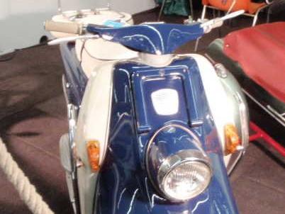 Blau-weißer Roller mit Logo und Scheinwerfer in einer Ausstellung, davor eine Absperrung aus Seil.