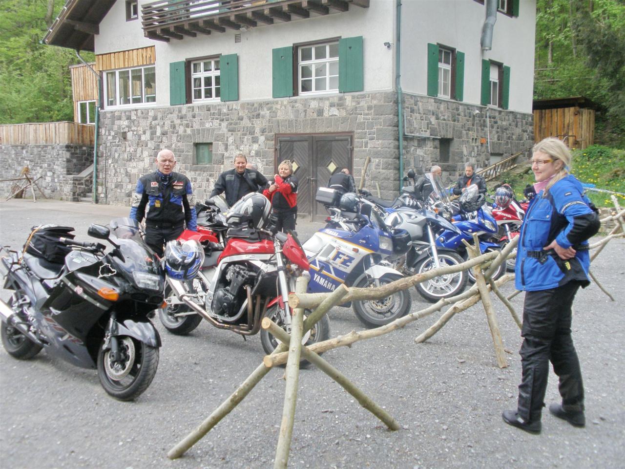 Mehrere Motorräder stehen vor einem steinernen Haus mit grünen Fensterläden, daneben stehen Menschen in Motorradkleidung.